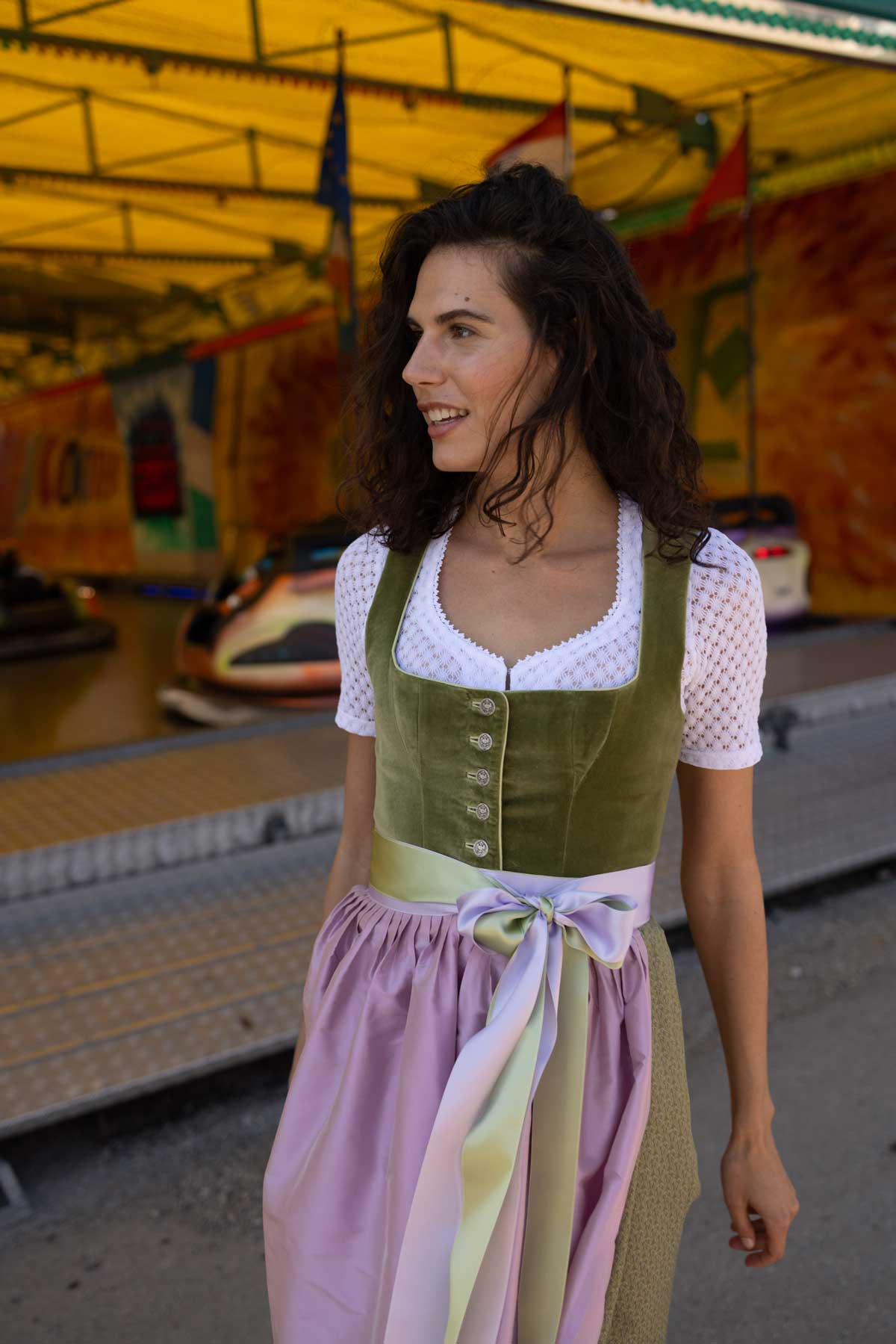 Frau im hellgrünen Samtdirndl mit silberner Knopfleiste, kombiniert mit weißer Dirndlbluse und rosafarbener Seidenschürze mit farblich abgestimmter Schleife – frischer Trachtenlook vor einem bunten Autoscooter.