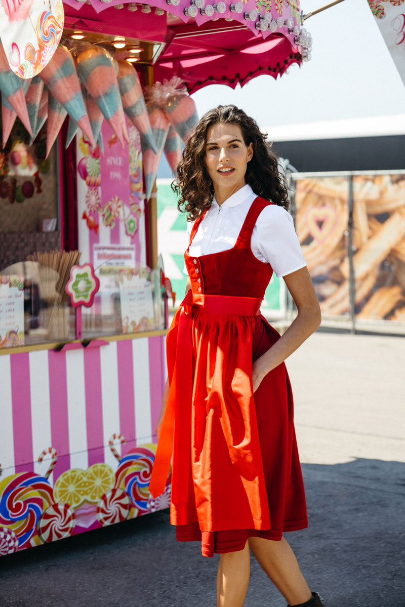 Frau in leuchtend rotem Samtdirndl mit Knopfleiste und farblich passender Schürze, kombiniert mit weißer Dirndlbluse – fröhlicher Auftritt vor einem nostalgischen Süßigkeitenstand mit Zuckerwatte.