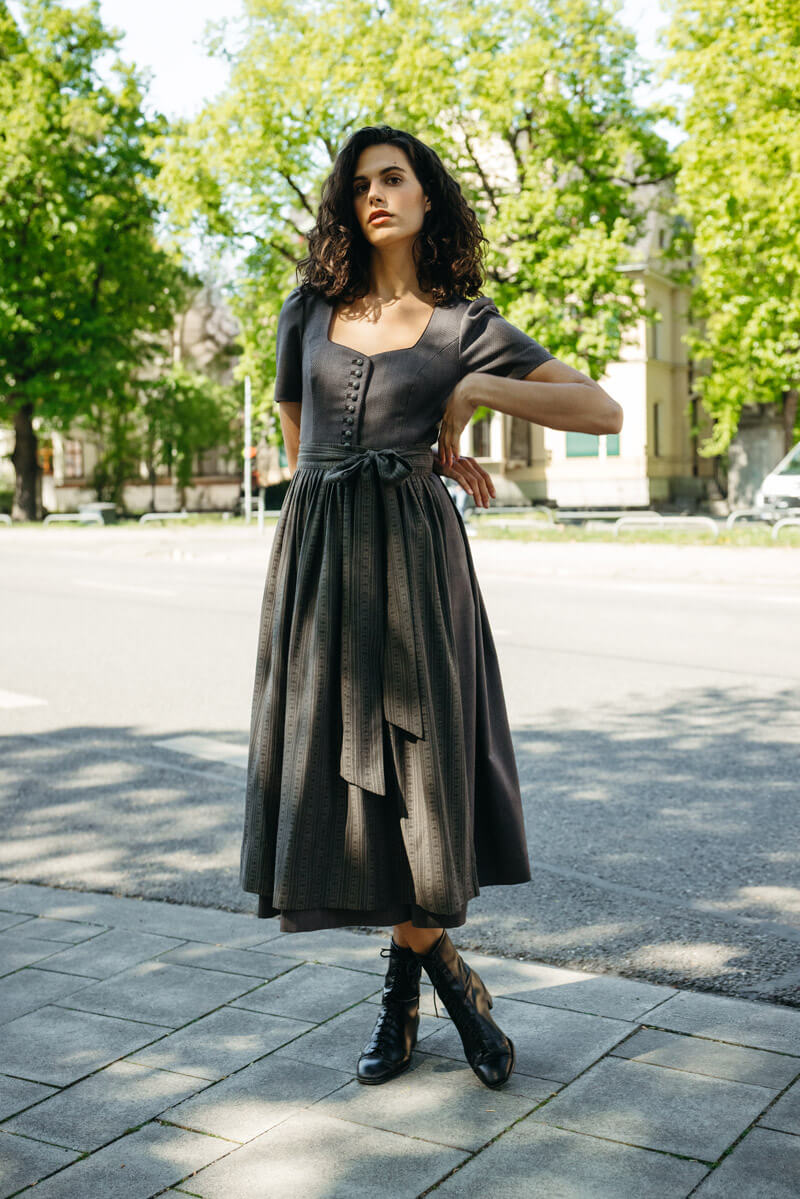 Frau in modernem Dirndl in Taupe mit halblangen Ärmeln, stoffbezogenen Knöpfen und gemusterter Schürze mit breiter Schleife – eleganter Trachtenlook auf städtischem Gehweg unter grünem Blätterdach.