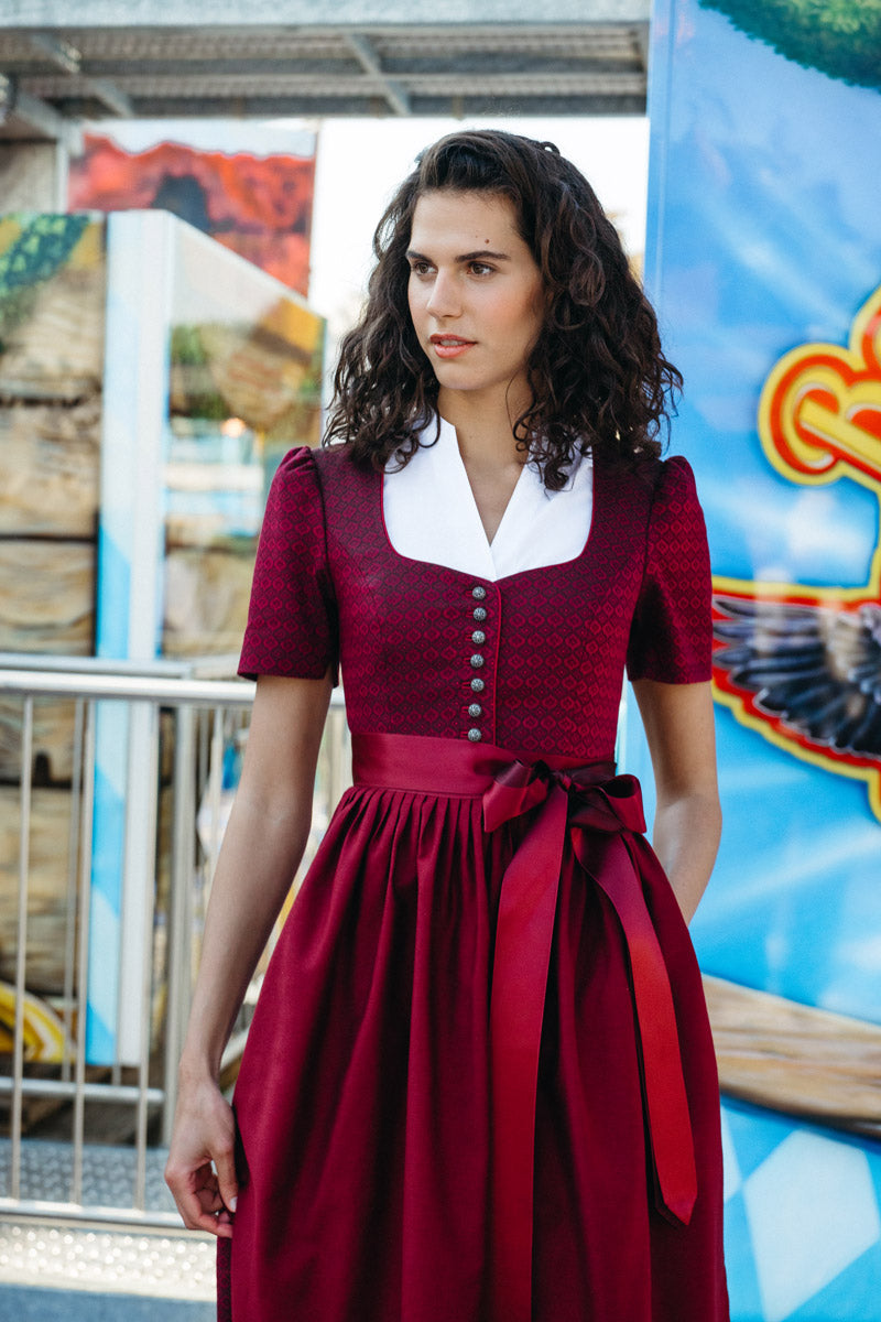 Frau in weinrotem Dirndl mit dezentem Muster, silberner Knopfleiste und tonaler Schürze mit breiter Schleife, kombiniert mit weißer Bluse – eleganter Auftritt vor bunter Jahrmarkt-Kulisse.
