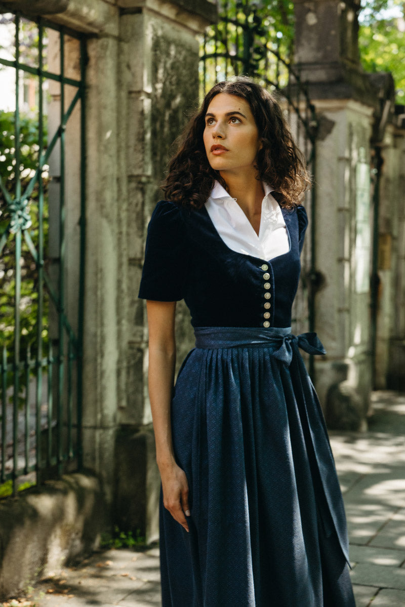 Frau im eleganten dunkelblauen Samtdirndl mit klassischem Ausschnitt, silberner Knopfleiste und gemusterter Baumwollschürze – stilvoller Look vor historischem Steinzaun im Schatten alter Bäume.