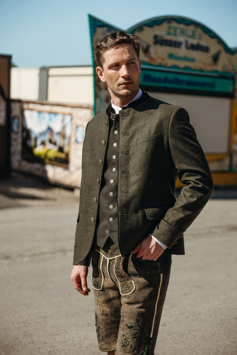 Mann in olivbraunem Trachtenjanker mit Stehkragen und gemusterter Weste, kombiniert mit bestickter Lederhose – traditioneller Look im Sonnenschein vor bayerischem Festzelt-Hintergrund.