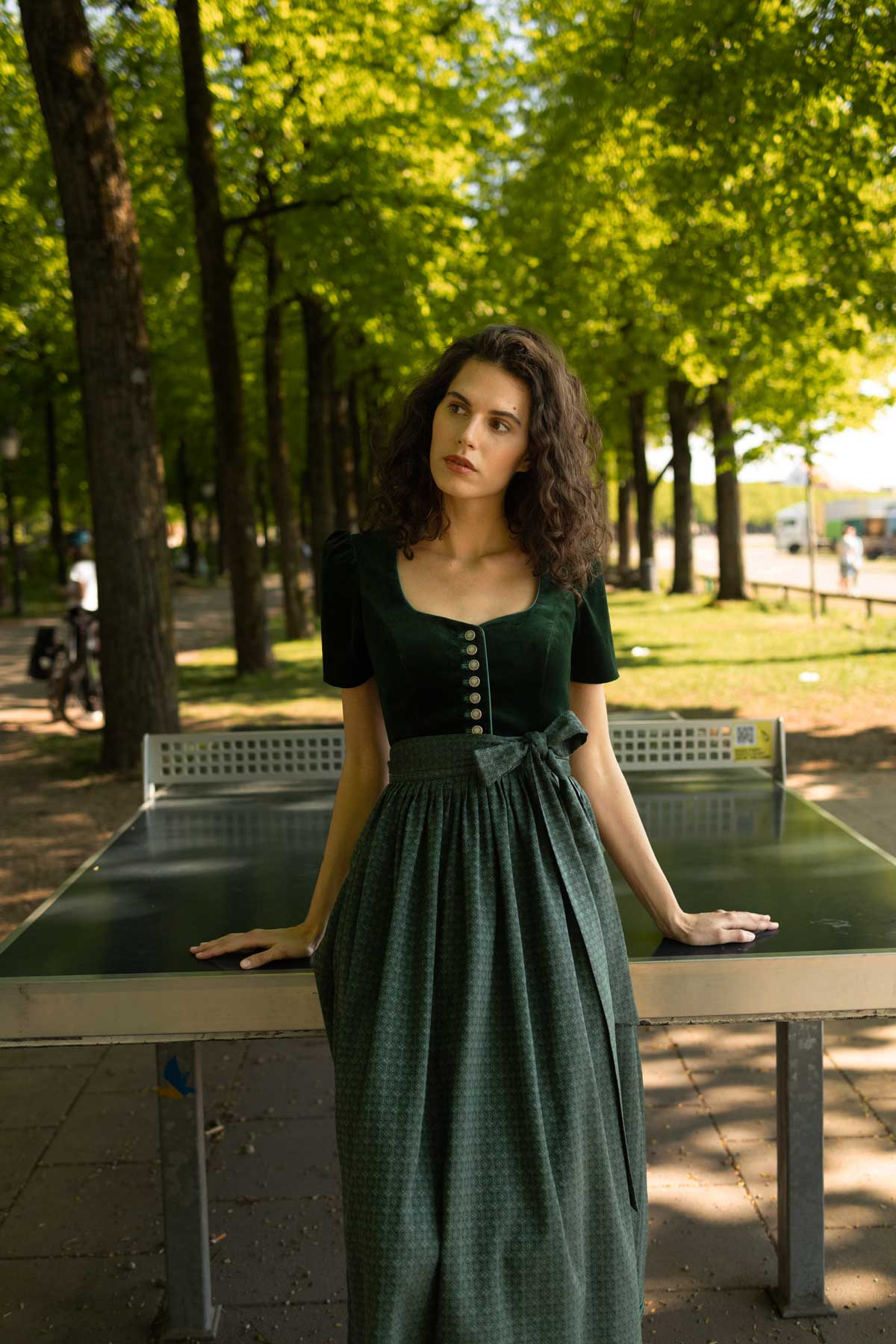 Frau in elegantem dunkelgrünem Samtdirndl mit Knopfleiste und gemusterter Baumwollschürze, stehend an einer Tischtennisplatte im schattigen Park – stilvoller Wiesn-Look mit natürlicher Eleganz.