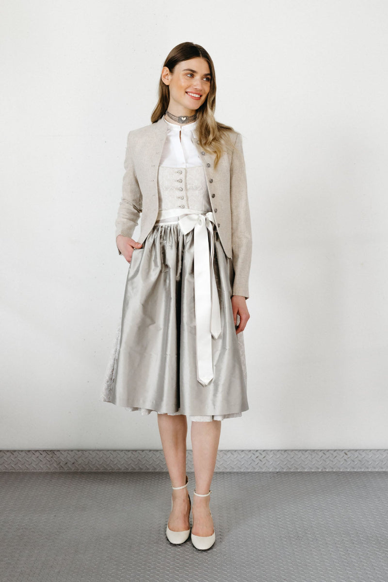 Frau trägt ein silbergraues Dirndl mit schimmerndem Rock und heller Schleife, kombiniert mit einer cremefarbenen Dirndlbluse und einem hellen Trachtenjanker; dazu beige Pumps, vor neutralem Studiohintergrund.