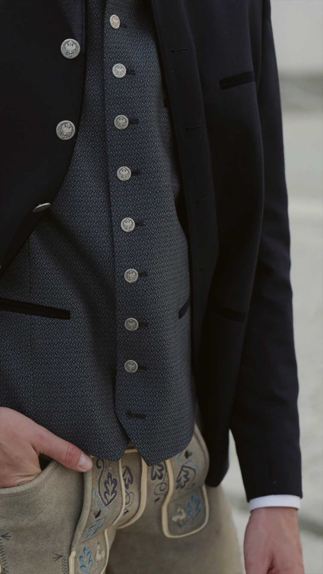 Trachtenoutfit mit dunkelblauem Janker, gemusterter Trachtenweste mit silbernen Münzknöpfen und heller Lederhose mit blauer Stickerei.