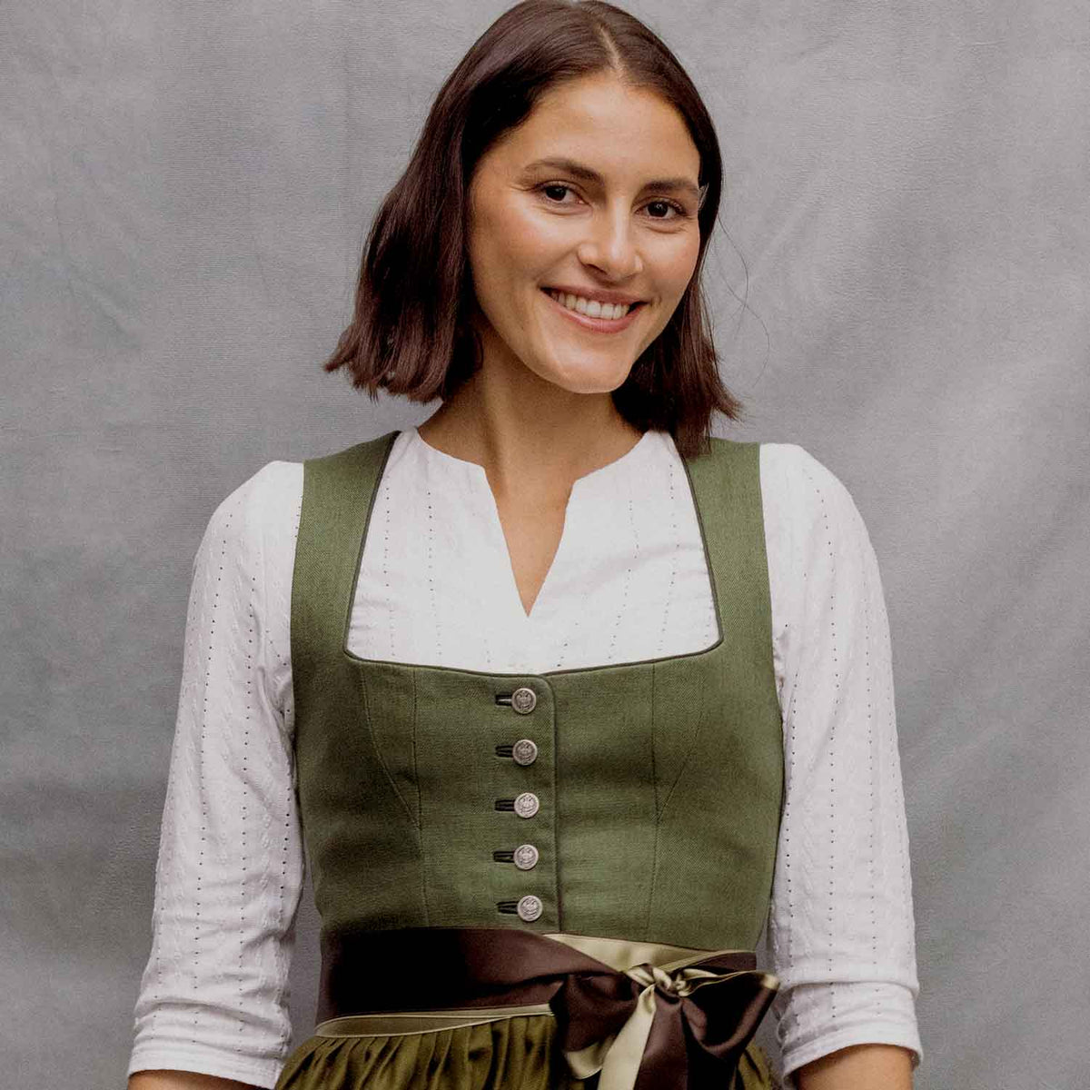 Junge Frau mit dunkelbraunem Bob trägt ein olivgrünes Dirndl mit eckigem Ausschnitt, silbernen Knöpfen und zweifarbiger Schleife in Braun und Champagner über einer weißen Dreiviertelarm-Bluse. Sie lächelt vor einem hellgrauen, strukturierten Studiohintergrund in die Kamera.
