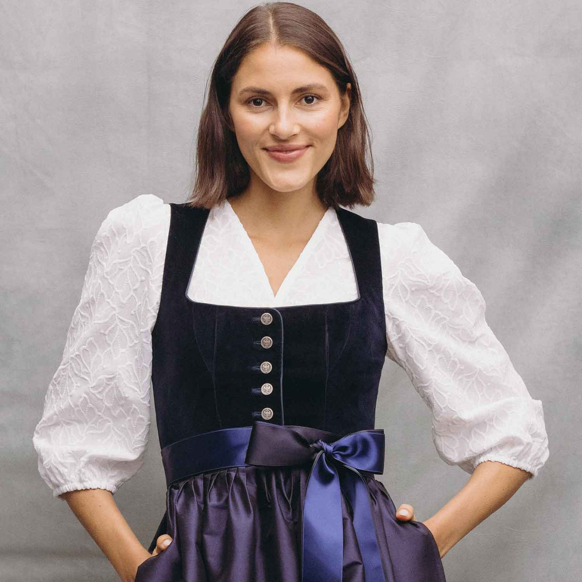 Junge Frau mit dunkelbraunem Bob trägt ein dunkelblaues Samtdirndl mit eckigem Ausschnitt, silbernen Knöpfen und zweifarbiger Schleife in Dunkelblau und Royalblau über einer weißen Dirndlbluse mit floraler Struktur und Puffärmeln. Sie steht lächelnd vor einem hellgrauen, strukturierten Studiohintergrund.