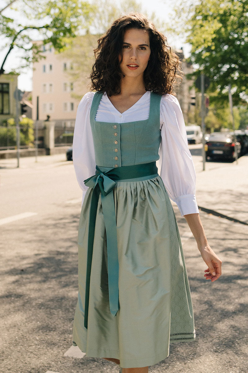 Model trägt Leinendirndl in Salbeigrün mit Seidenschürze und weißer Dirndlbluse – modernes Midi Dirndl von Amsel Fashion im sommerlichen Stadtlook
