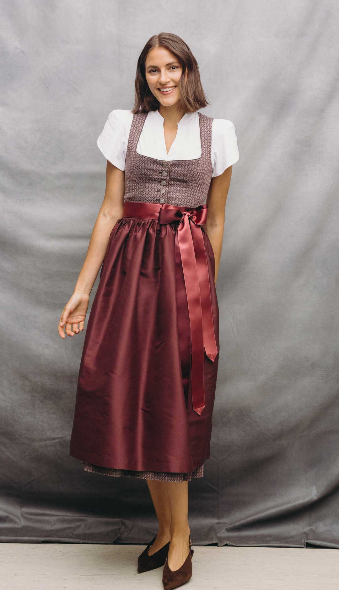 Model trägt ein rotes Dirndl mit gemustertem Mieder, weißer Dirndlbluse und roter Schürze mit Satinbändern vor grauem Studiohintergrund.