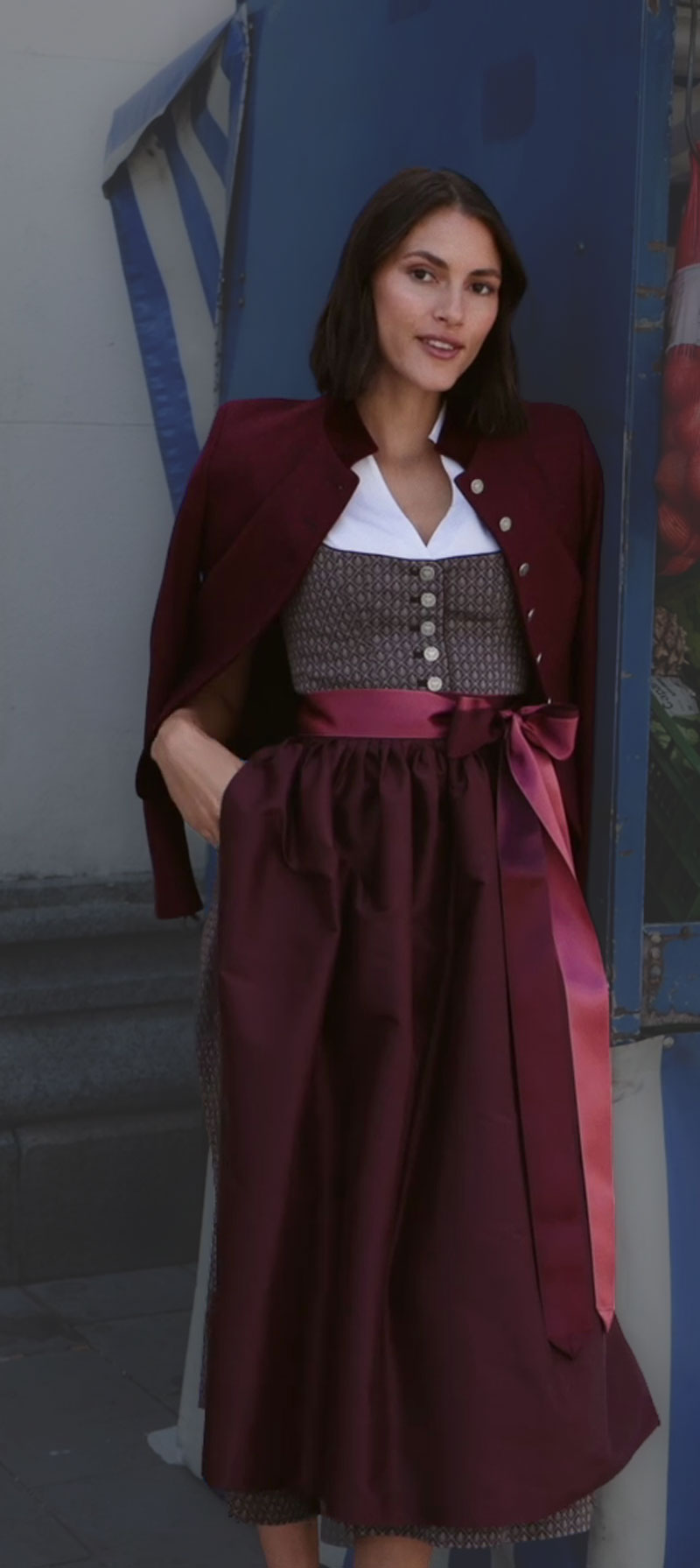 Frau im bordeauxfarbenen Dirndl mit gemustertem Mieder, weißer Bluse und farblich abgestimmter Seidenschürze, kombiniert mit bordeauxfarbenem Janker.