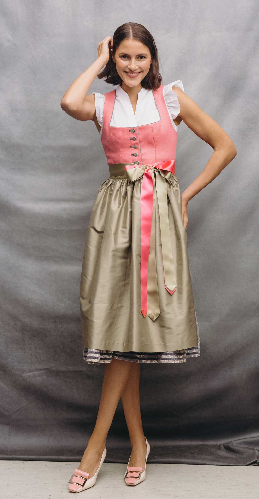 Model trägt ein rosa Dirndl mit heller Schürze, zweifarbigen Satinbändern und weißer Dirndlbluse vor grauem Studiohintergrund.