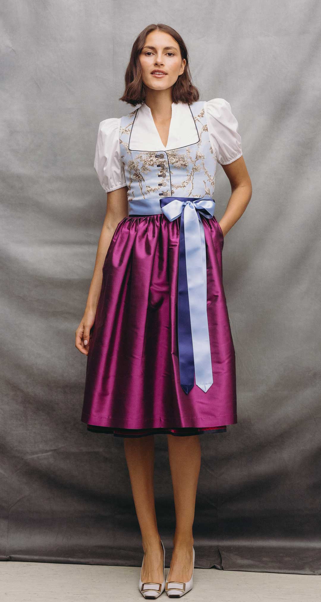 Model trägt ein Dirndl mit hellblauem Toile-de-Jouy-Mieder, pinkfarbener Schürze, zweifarbigen Satinbändern und weißer Dirndlbluse vor grauem Studiohintergrund.