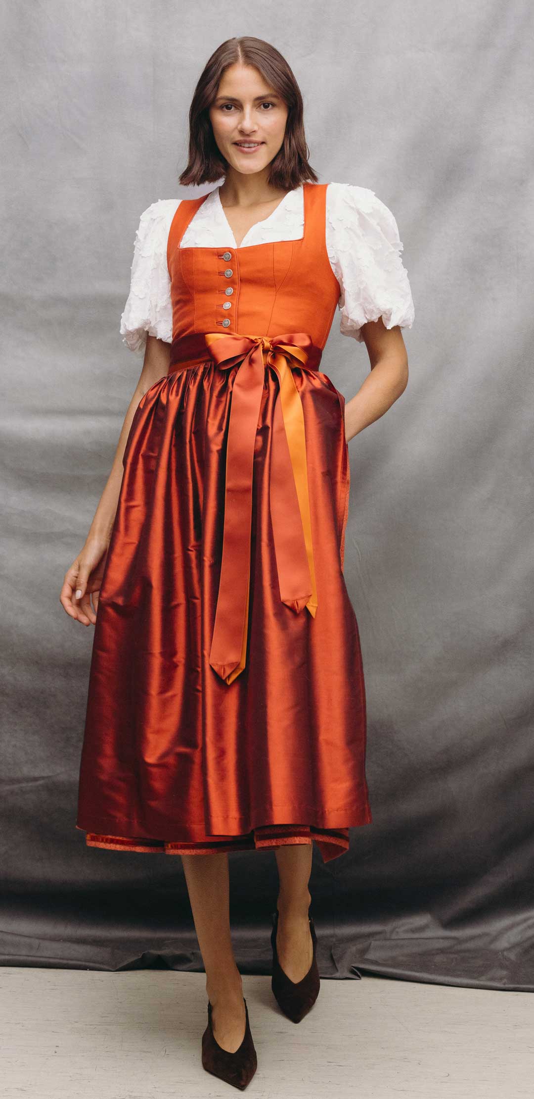 Frau trägt ein elegantes Dirndl in Orange mit rot-orange schimmerndem Rock, zweifarbigem Satinband und weißer Dirndlbluse mit strukturierten Puffärmeln.