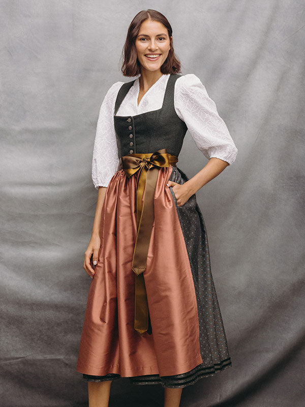 Frau im langen Dirndl mit grünem Mieder, weißer Bluse, roséfarbener Seidenschürze und brauner Schleife vor grauem Hintergrund.