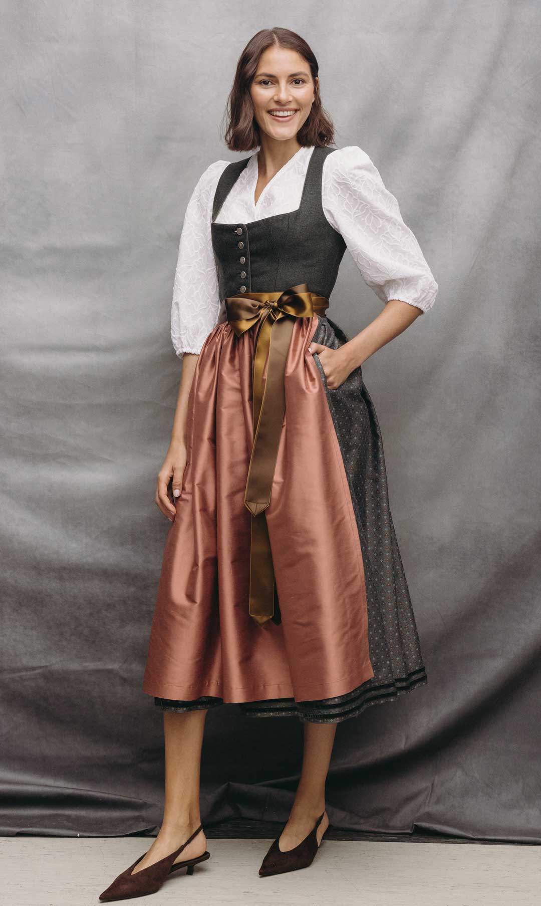 Model trägt ein dunkles Dirndl mit weißer Dirndlbluse, roséfarbener Schürze und braunen Satinbändern vor grauem Studiohintergrund.