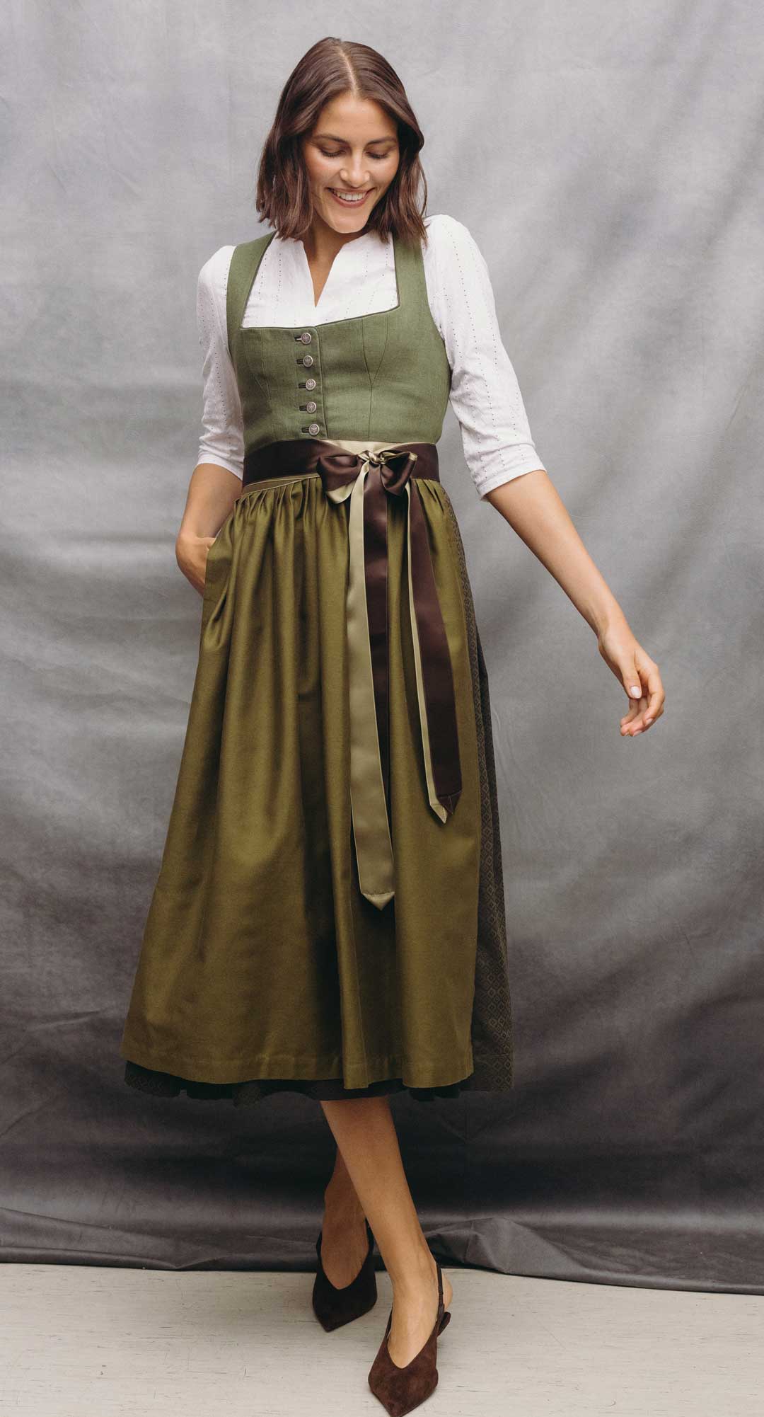 Model trägt ein olivgrünes Dirndl mit weißer Dirndlbluse, zweifarbigen Satinbändern und dunklen Pumps vor grauem Studiohintergrund.