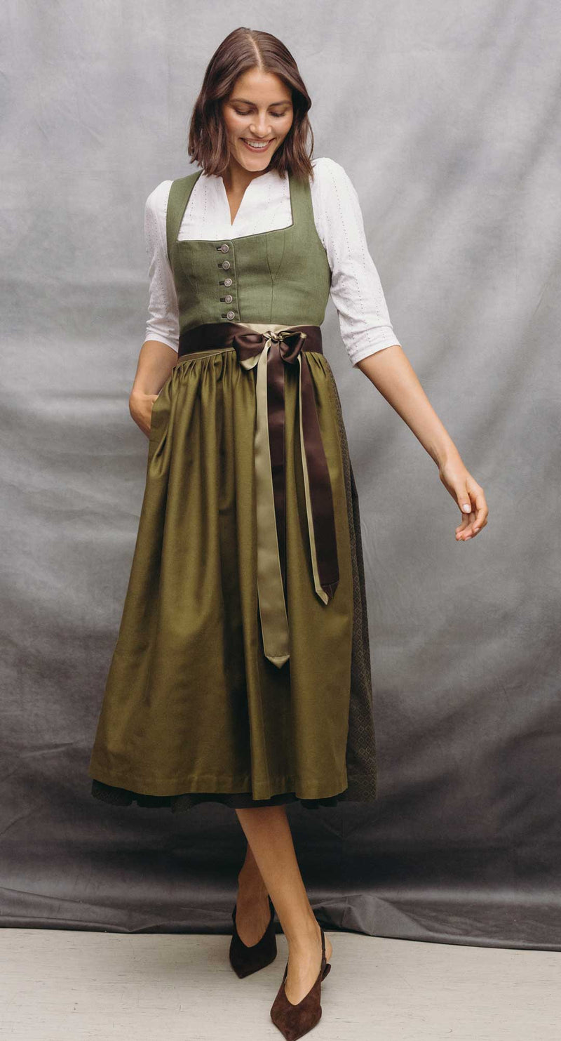 Model trägt ein olivgrünes Dirndl mit weißer Dirndlbluse, zweifarbigen Satinbändern und dunklen Pumps vor grauem Studiohintergrund.