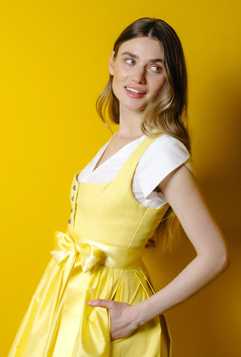 Model trägt gelbes Dirndl mit weißer Dirndlbluse vor gelbem Hintergrund – frischer, sommerlicher Trachtenlook von Amsel
