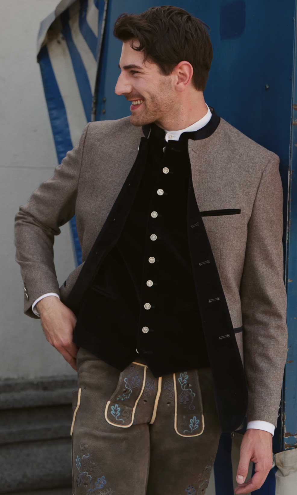 Trachtenoutfit mit greigefarbenem Janker, schwarzer Samtweste mit Münzknöpfen und brauner Hirschlederhose mit blauer Stickerei.