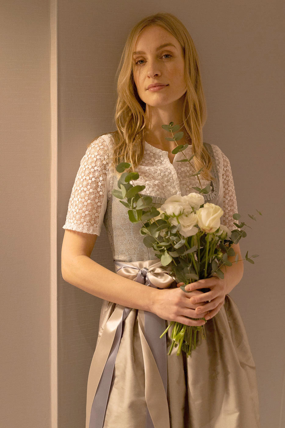 Braut in elegantem cremefarbenen Dirndl mit Spitzenbluse und Blumenstrauß, Hochzeitsdirndl von Amsel Fashion.