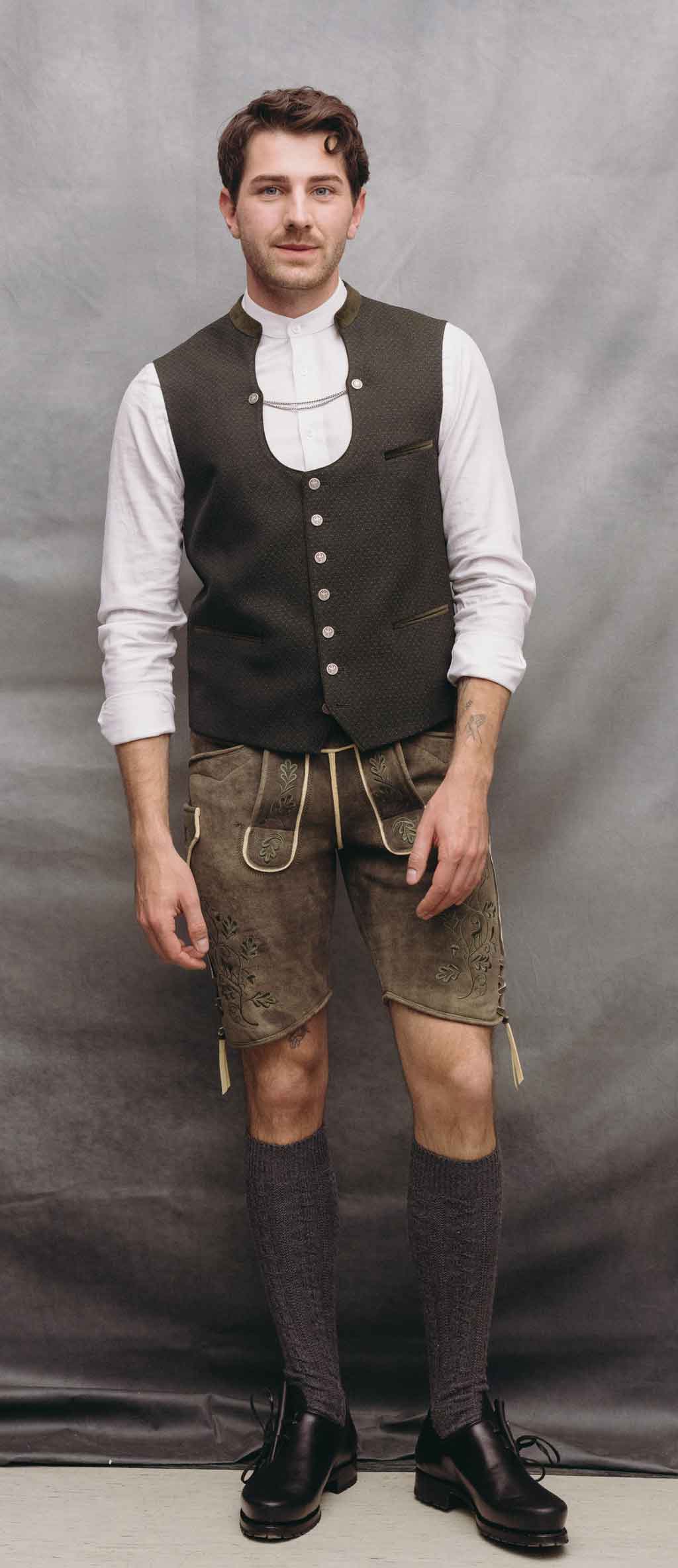 Mann mit dunkelolivfarbener Trachtenweste, weißem Stehkragenhemd und hellbrauner Hirschlederhose vor hellgrauem Studiohintergrund.