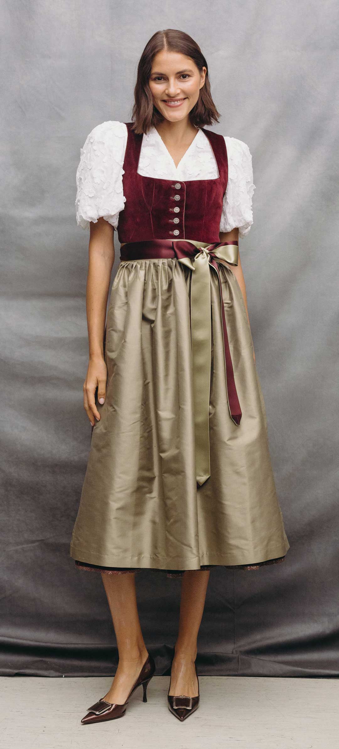 Bordeauxrotes Samtdirndl mit eckigem Ausschnitt, weißer Dirndlbluse mit floraler Struktur und Puffärmeln sowie schimmernder Doupionseiden-Schürze in Taupe mit zweifarbigem Satinband in Bordeaux und Taupe – elegantes Premium-Dirndl für Wiesn, Hochzeit und festliche Anlässe.