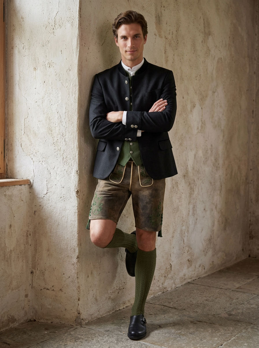 Eleganter Trachtenlook für Herren mit schwarzem Janker, grüner Trachtenweste, weißem Stehkragenhemd und Hirschlederhose mit grüner Stickerei.