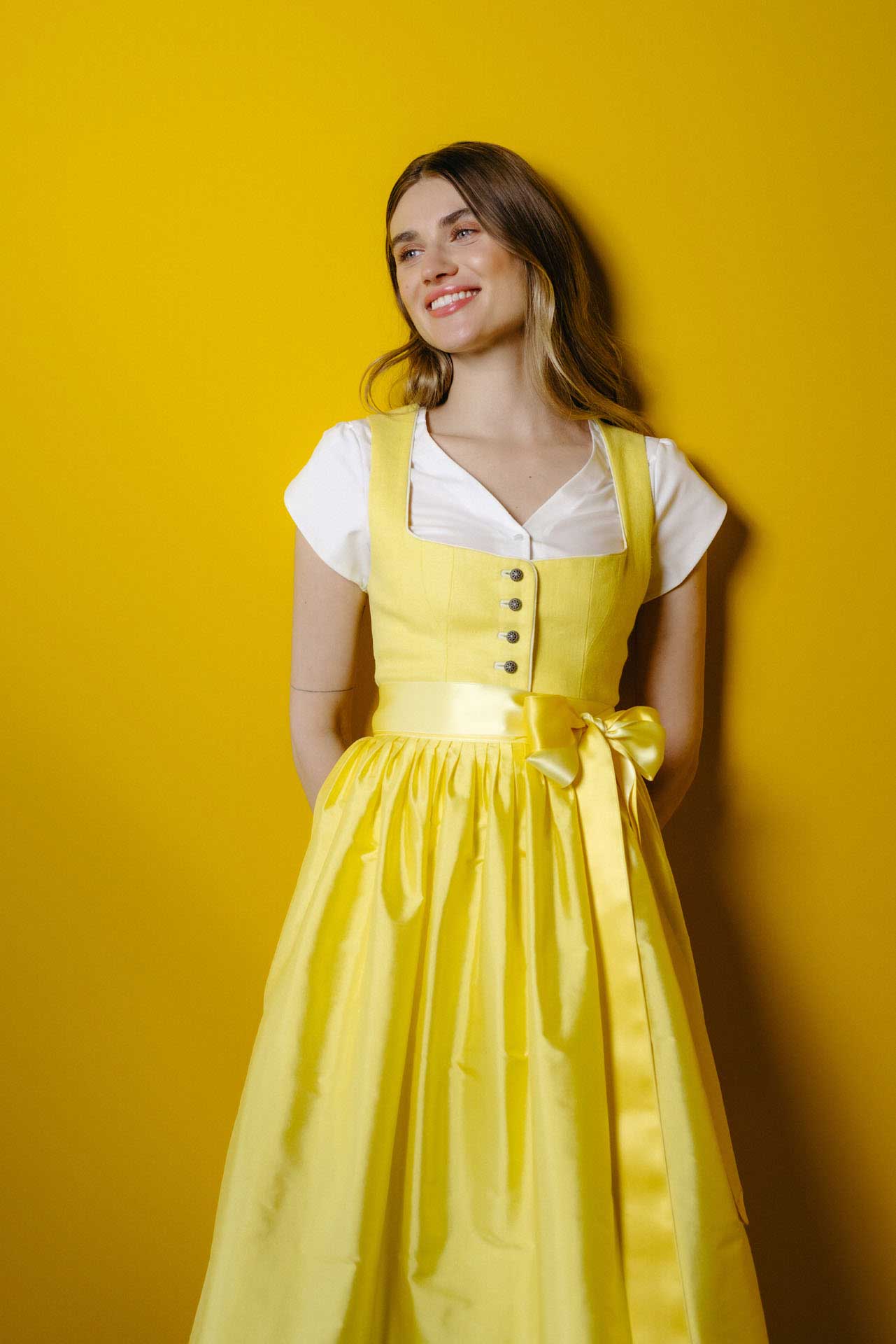 Model trägt ein leuchtend gelbes Dirndl mit klassischem Ausschnitt, silbernen Knöpfen und breitem Satinband, kombiniert mit weißer Dirndlbluse, vor gelbem Hintergrund – moderner, frischer Look.