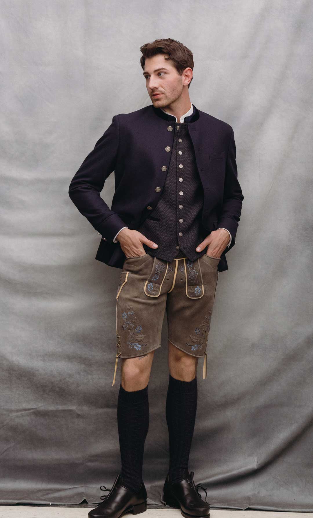 Trachtenoutfit mit dunkelblauem Janker, dunkel gemusterter Trachtenweste mit Münzknöpfen und brauner Lederhose aus sämisch gegerbtem Ziegenleder mit blauer Stickerei.