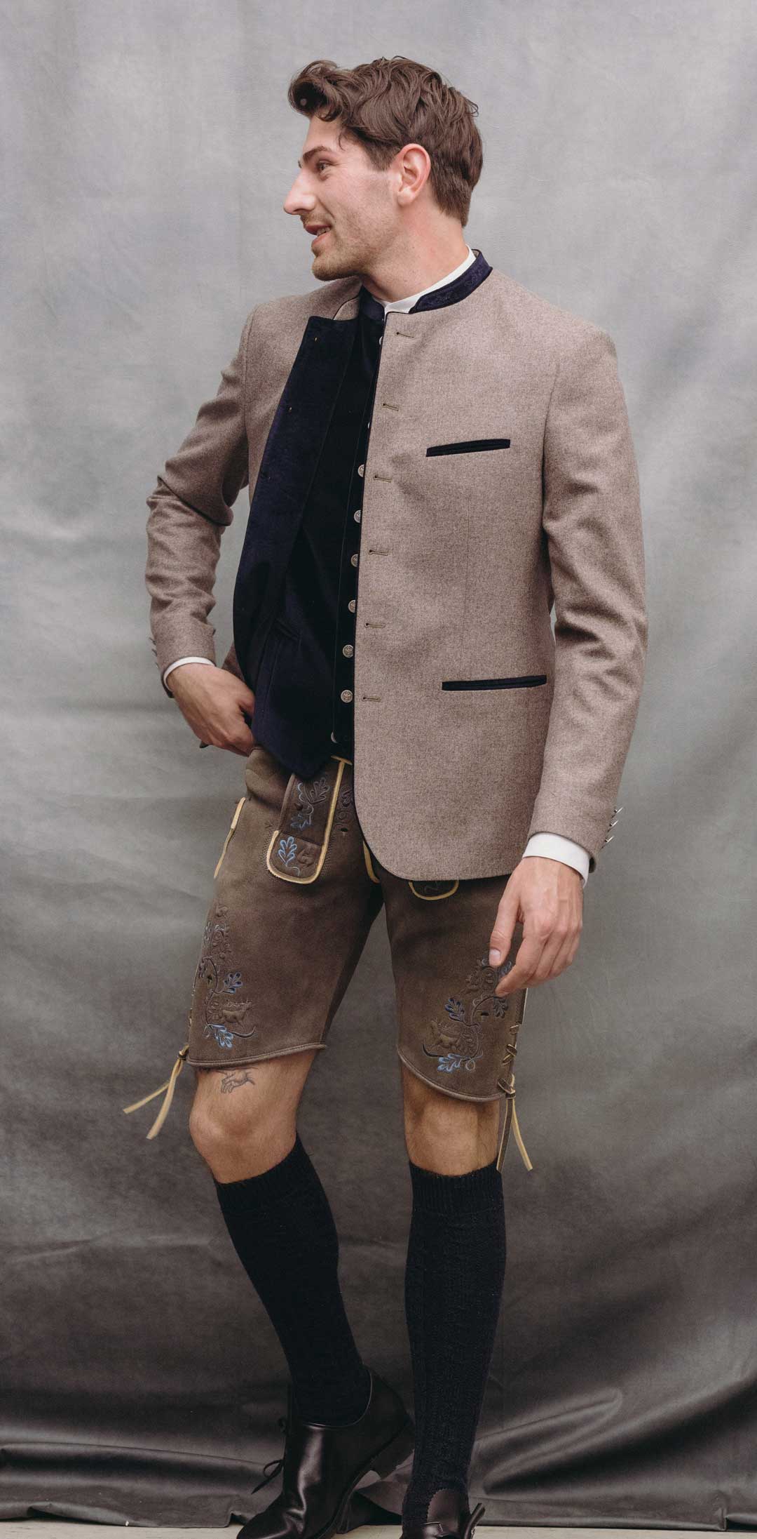Trachtenoutfit mit greigefarbenem Janker, dunkelblauer Trachtenweste mit Münzknöpfen und Hirschlederhose in Braun mit blauer Stickerei.
