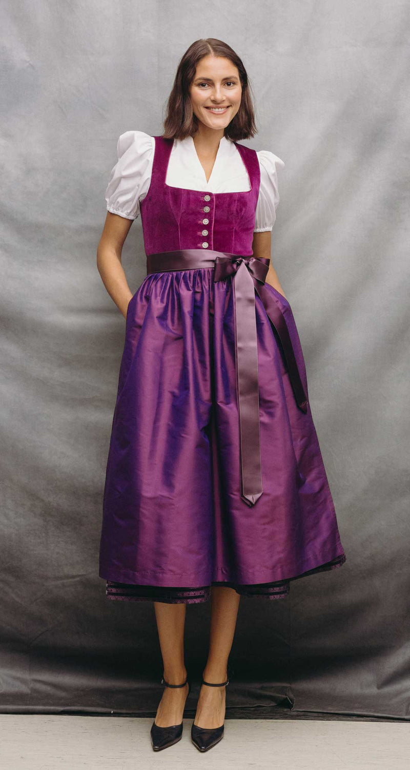 Frau trägt ein elegantes Samtdirndl in Beere mit violett schimmerndem Rock, dunkler Satinschürze und weißer Dirndlbluse mit Puffärmeln.