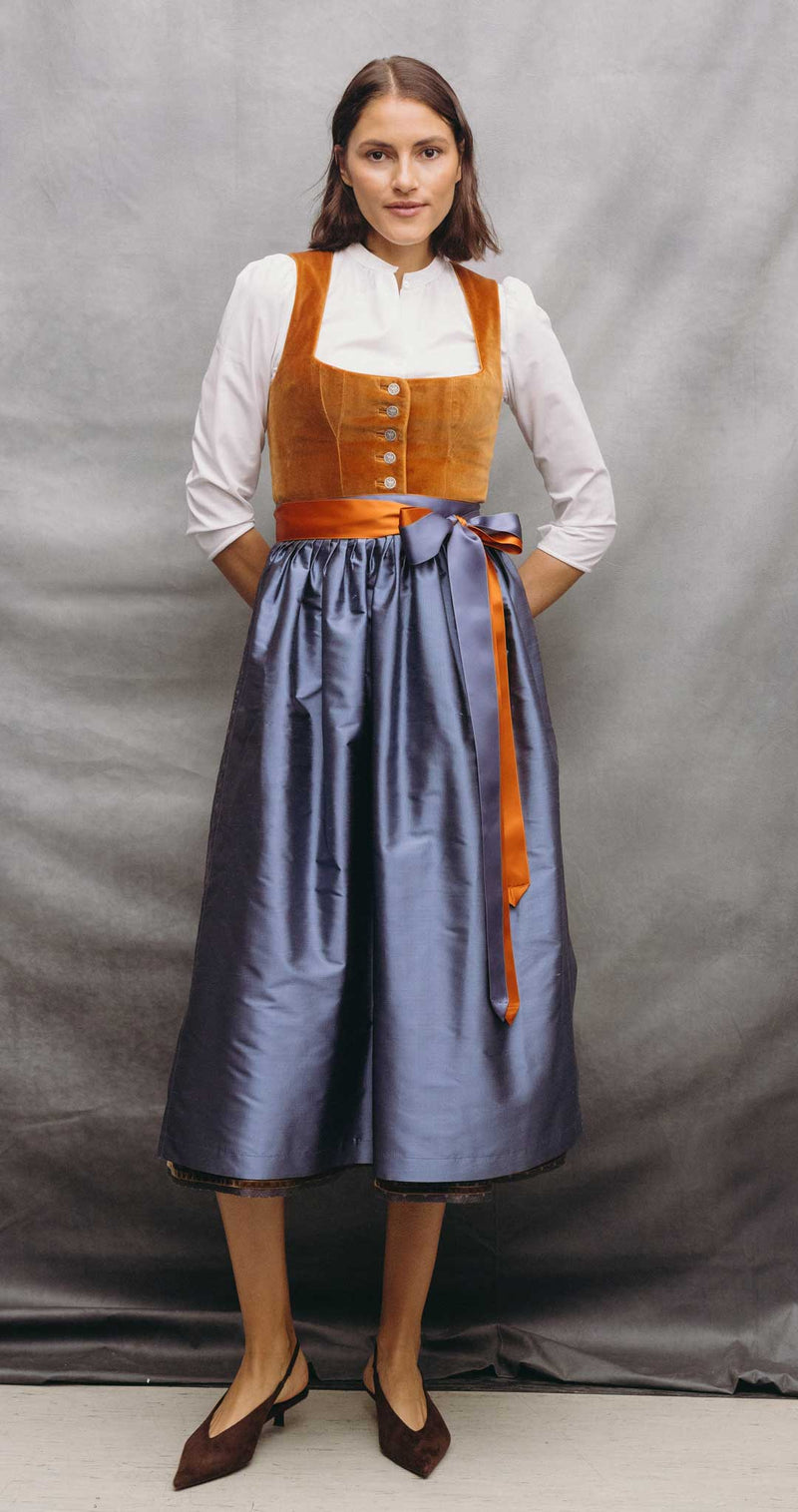 Frau trägt ein elegantes Samtdirndl in Karamell mit langem, blaugrau schimmerndem Rock, zweifarbigem Satinband und weißer Dirndlbluse.