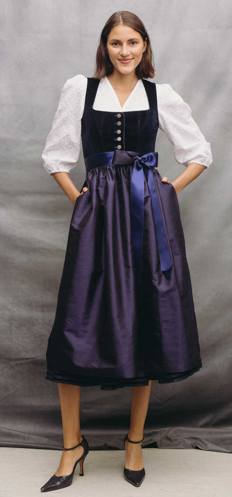 Frau trägt ein elegantes Samtdirndl in Dunkelblau mit violett schimmerndem Rock, zweifarbigem Satinband und weißer Dirndlbluse mit Stickerei.