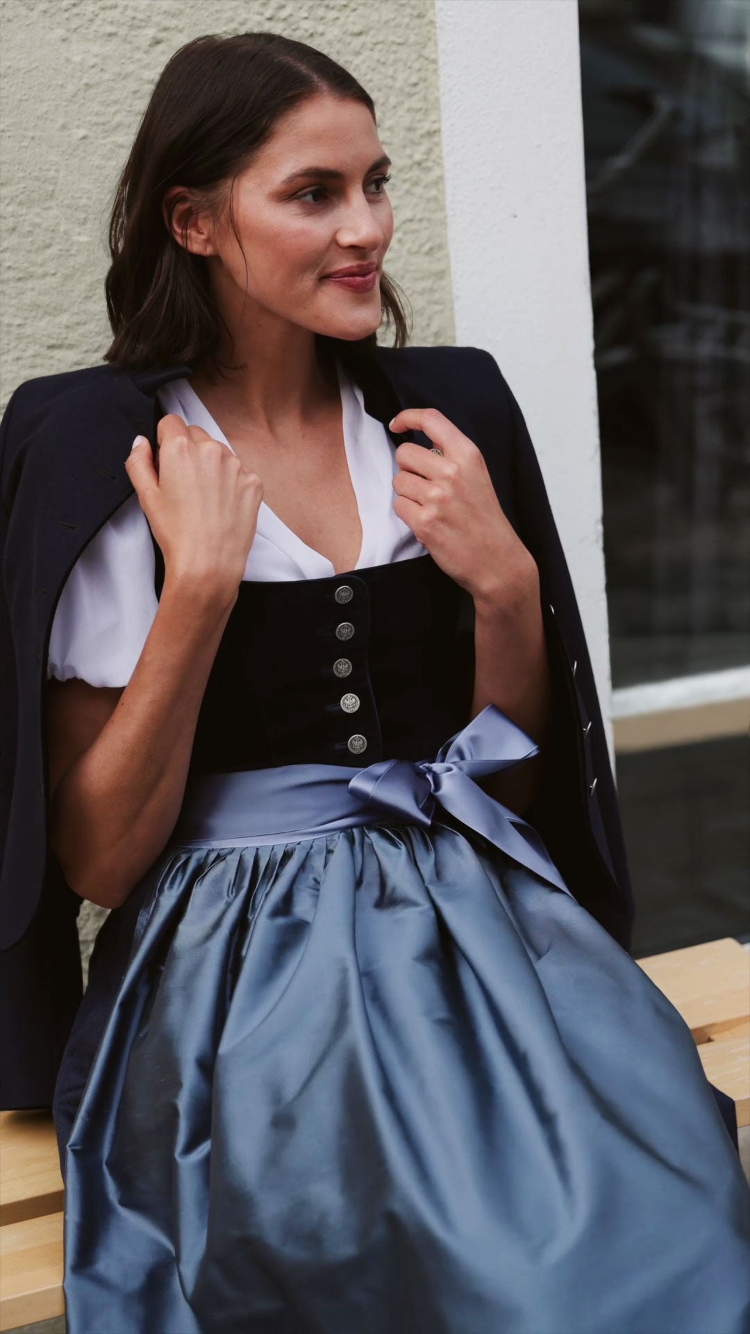Frau im blauen Dirndl Olivia mit hellblauer Seidenschürze und weißer Bluse, kombiniert mit dunkelblauem Janker über den Schultern.