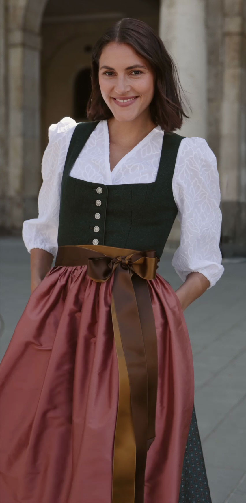 Dunkelgrünes Samtdirndl mit eckigem Ausschnitt und silbernen Knöpfen, kombiniert mit einer weißen Dirndlbluse mit dezentem floralem Muster und Puffärmeln. Dazu eine glänzende altrosafarbene Seidenschürze mit breitem schokoladenbraunem Schleifenband. Das Model steht lächelnd vor einer steinernen Kulisse.