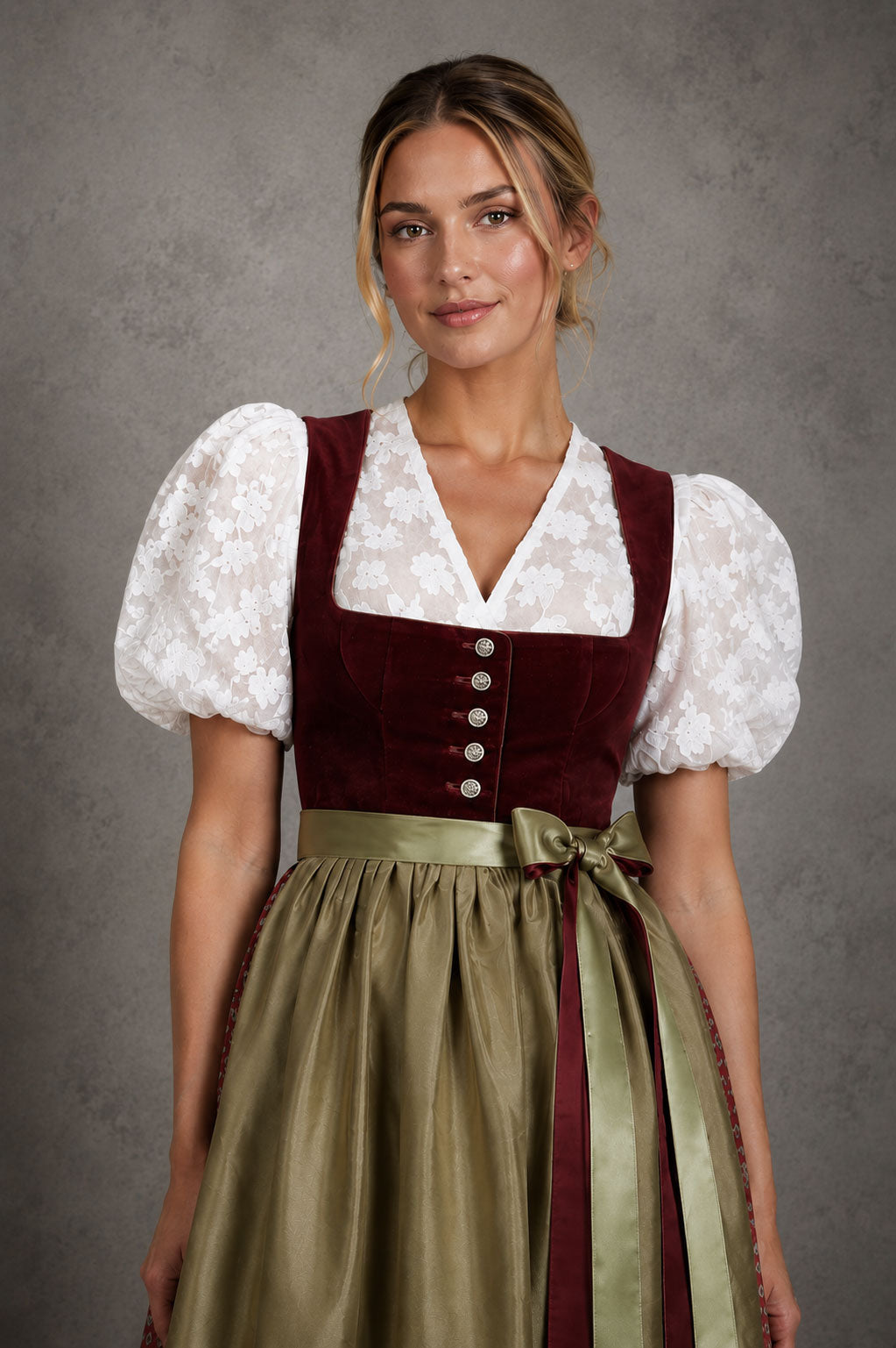 Blonde Frau im bordeauxfarbenen Samtdirndl mit weißer Puffarmbluse und olivgrüner Schürze vor einer steingrauen Studio-Wand.