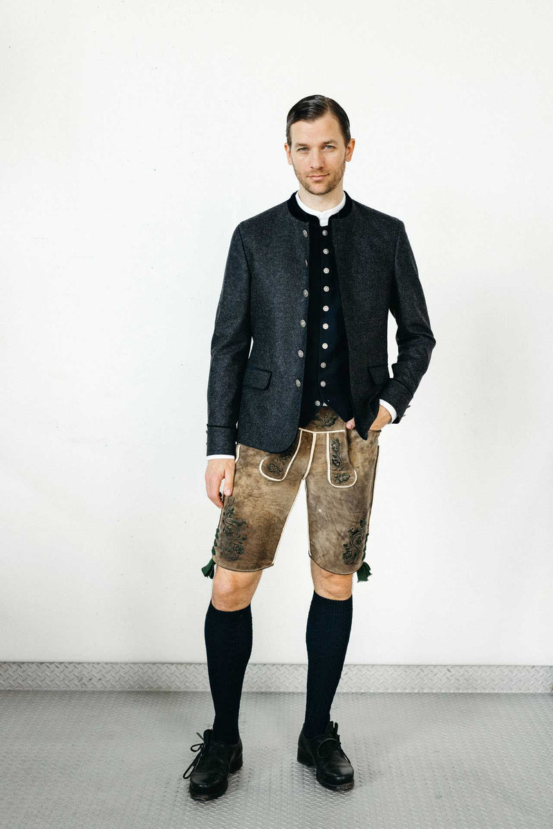 Mann in anthrazitfarbenem Trachtenjanker mit dunkelblauer Weste, weißem Stehkragenhemd und hellbrauner Lederhose vor hellem Studiohintergrund.