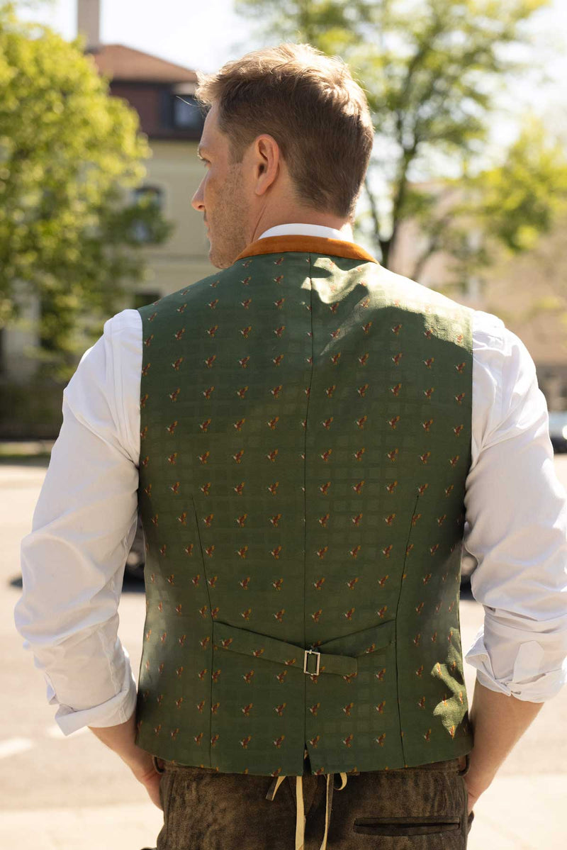 Rückansicht eines Mannes in weißem Hemd und grüner Weste mit dezentem Enten-Muster und orangefarbenem Kragen, kombiniert mit heller Lederhose – eleganter Trachtenstil mit liebevollen Details.