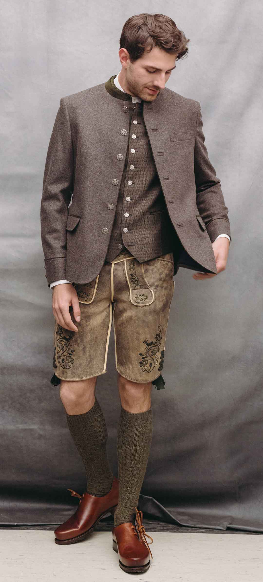 Mann in taupefarbenem Trachtenjanker mit gemusterter Weste und heller Lederhose vor grauem Studiohintergrund.