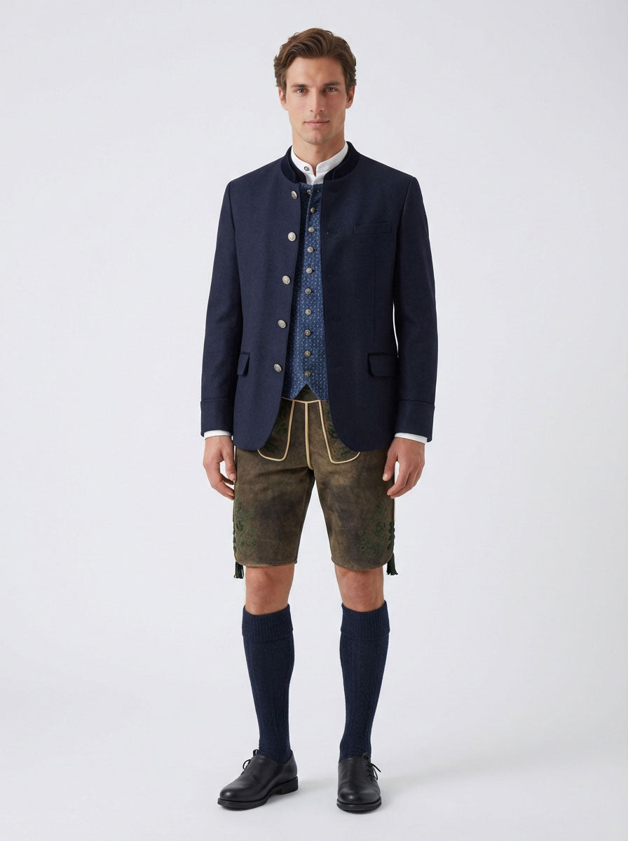 Männliches Model trägt eine traditionelle Lederhose mit blauer Trachtenweste, weißem Stehkragenhemd und einem dunkelblauen Janker über der Weste, kombiniert mit kniehohen Trachtensocken und schwarzen Haferlschuhen aus Leder vor neutralem Studiohintergrund.